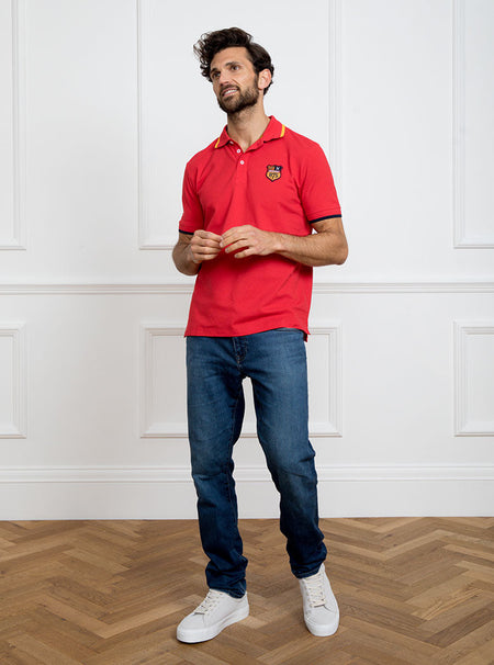 Man wearing red polo shirt in 100% cotton with Newmarket 875 crest embroidered on the front with navy sleeve trimming and gold collar trimming.